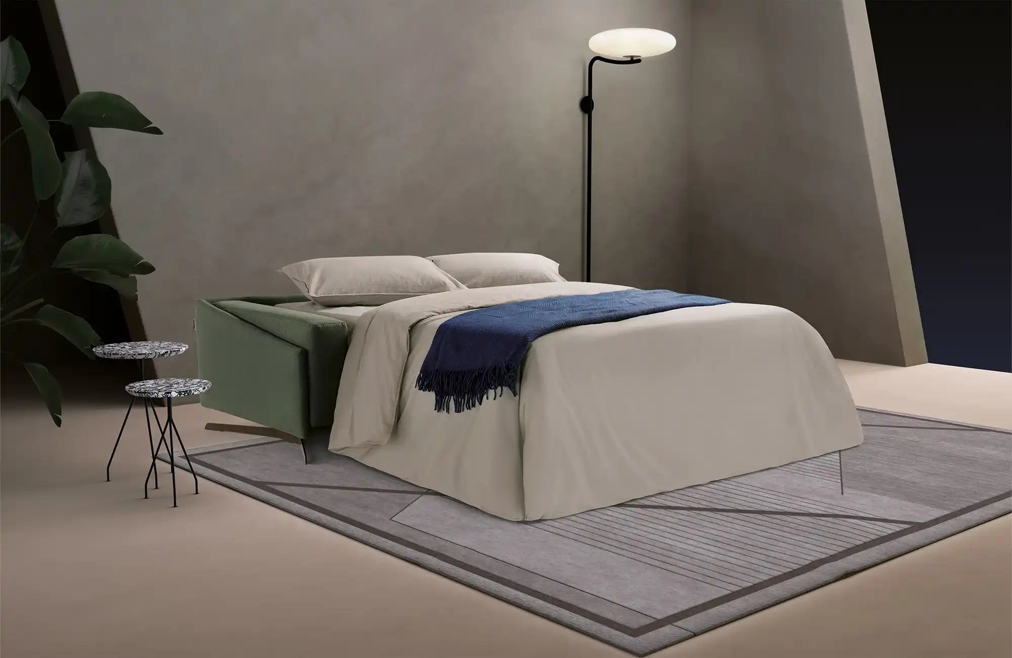 Ein modernes umwandelbares Sofabett mit einem salbeigrünen Rahmen, bedeckt mit hellfarbiger Bettwäsche und einer dunkelblauen Decke.